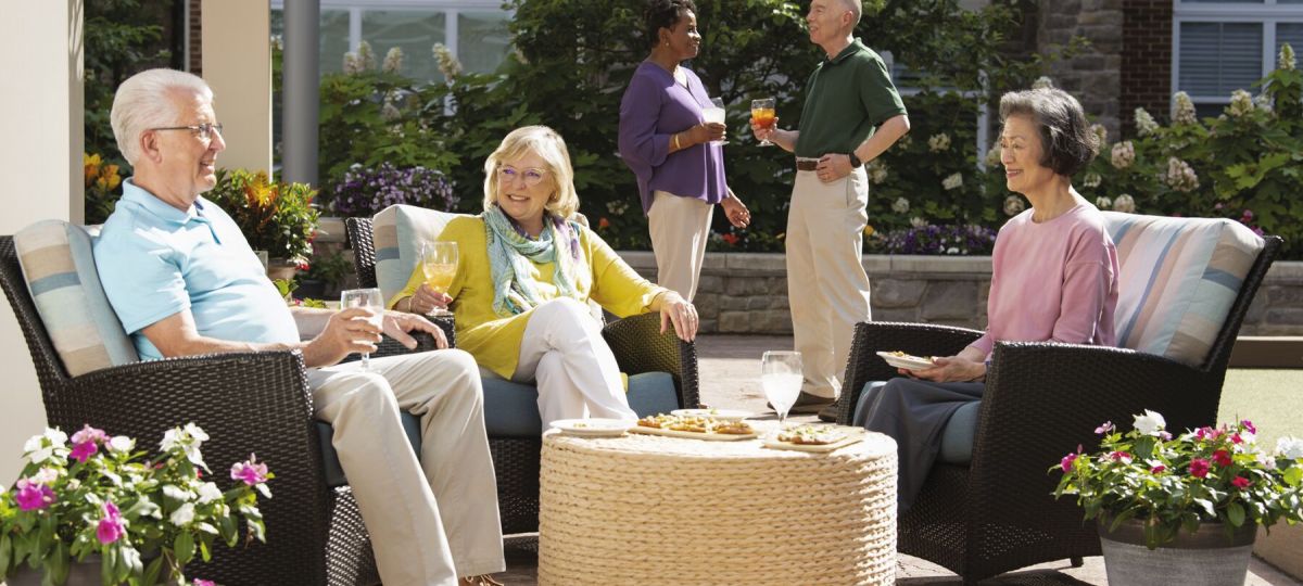 Group of older adults relaxing on an outdoor patio, sitting and standing together while enjoying drinks and conversation in a landscaped courtyard setting at an Erickson Senior Living community. 