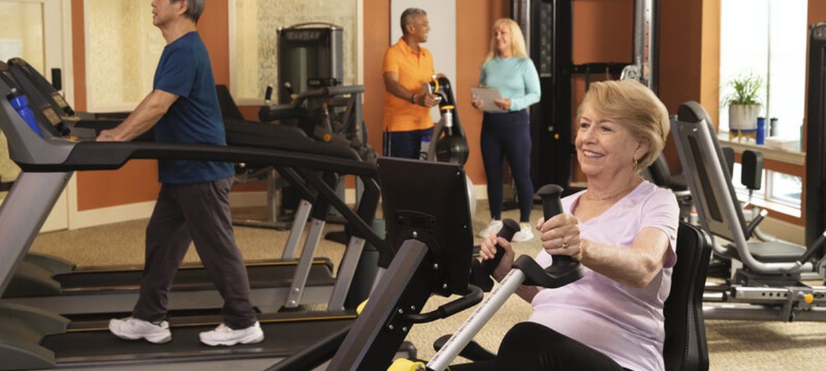 Older adults exercising in a senior living fitness center, including a woman using a rowing machine and a man walking on a treadmill, with staff assisting in the background.