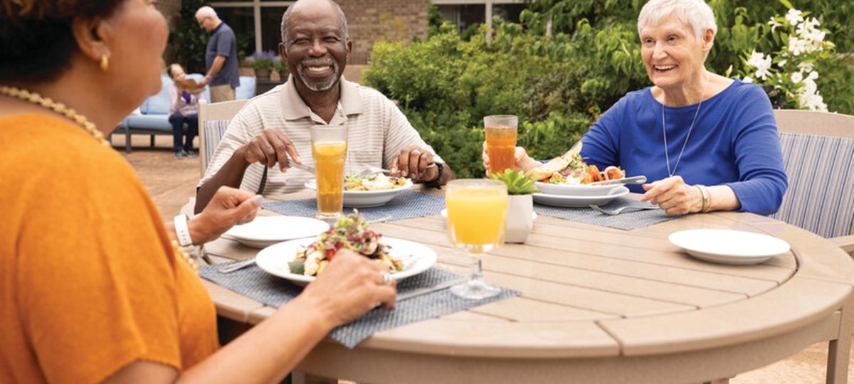 Three older adults enjoying a meal together at an outdoor patio table, smiling and talking in a senior living community setting.