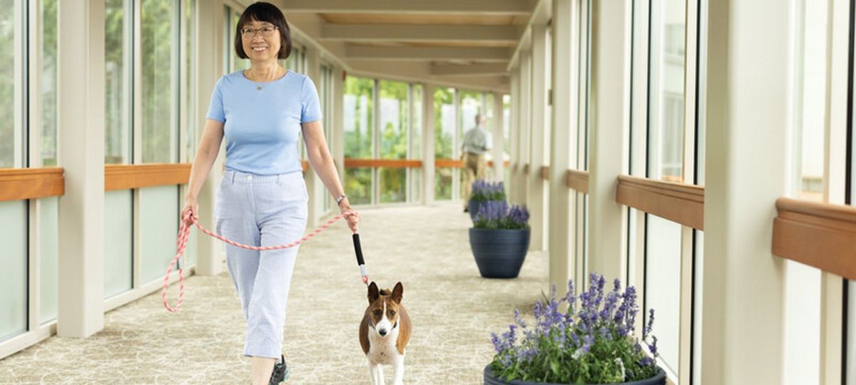 Older woman walking a small dog down a bright indoor hallway in a senior living community, with windows and potted plants along the corridor.