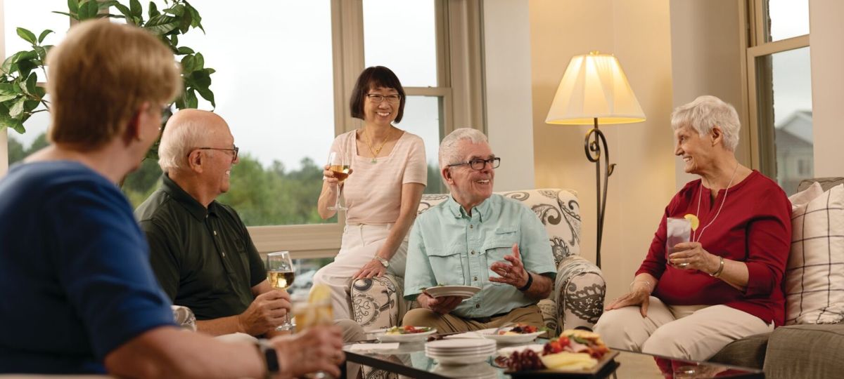 Group of older adults gathered in a bright living room, sitting together and enjoying drinks and conversation around a coffee table.