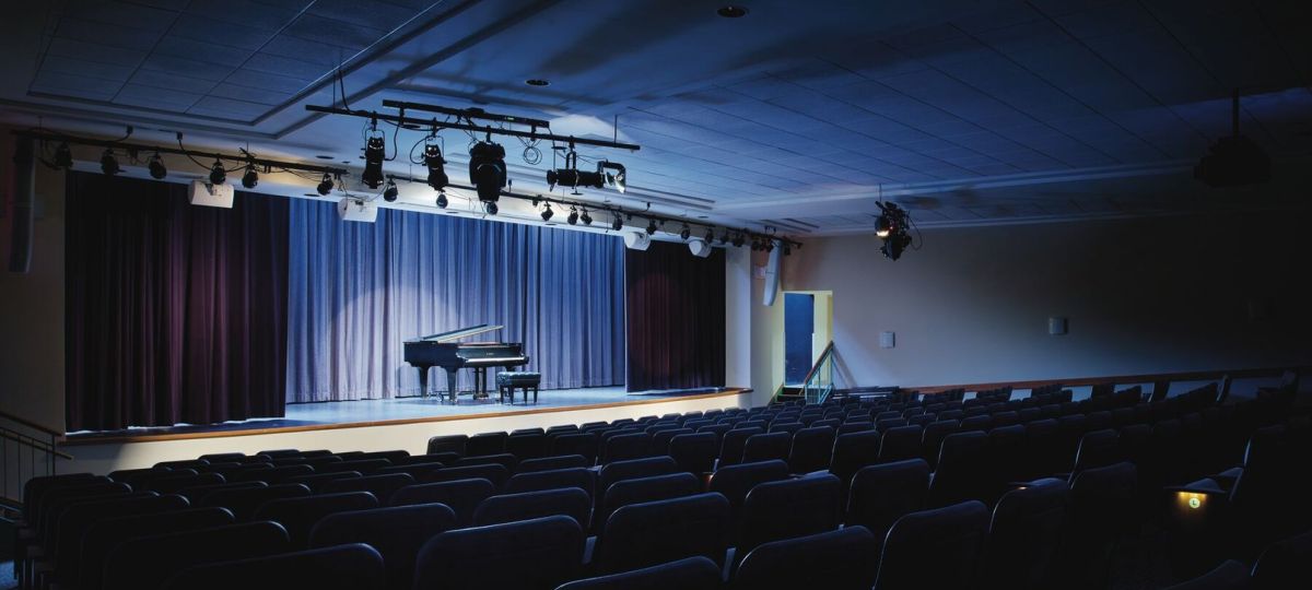 Empty auditorium with rows of seats facing a softly lit stage featuring a grand piano and closed curtains.