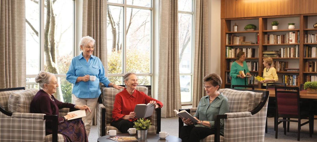 Group of older adults relaxing and reading together in a bright, cozy lounge with large windows and bookshelves, enjoying a social community setting.