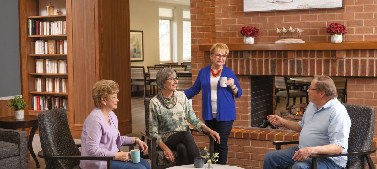 Four older adults sitting and chatting comfortably around a table by a fireplace in a cozy community lounge with bookshelves and warm decor.
