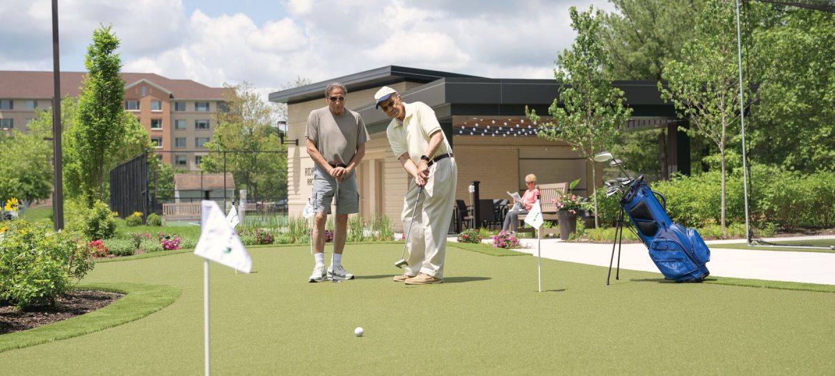 Two older men playing golf on a putting green in a landscaped outdoor area. 
