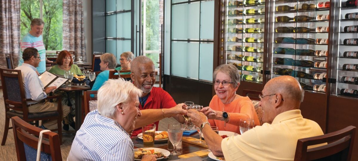 Group of older adults dining together at a restaurant table, smiling and toasting with drinks beside a wall of wine bottles.