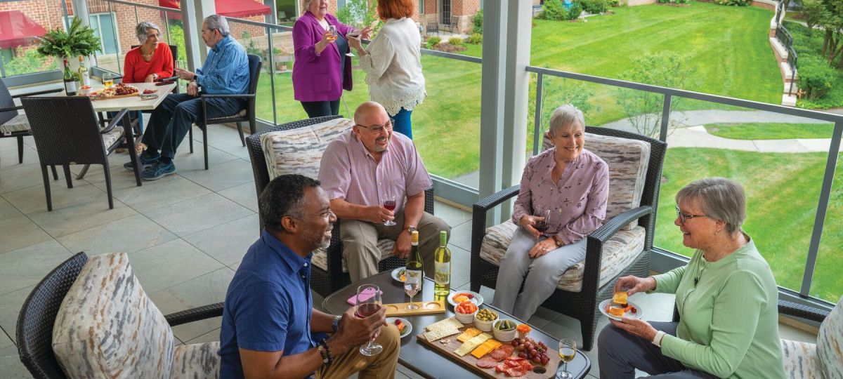 Group of older adults relaxing and socializing on a covered patio, enjoying drinks and appetizers together with a landscaped courtyard in the background.