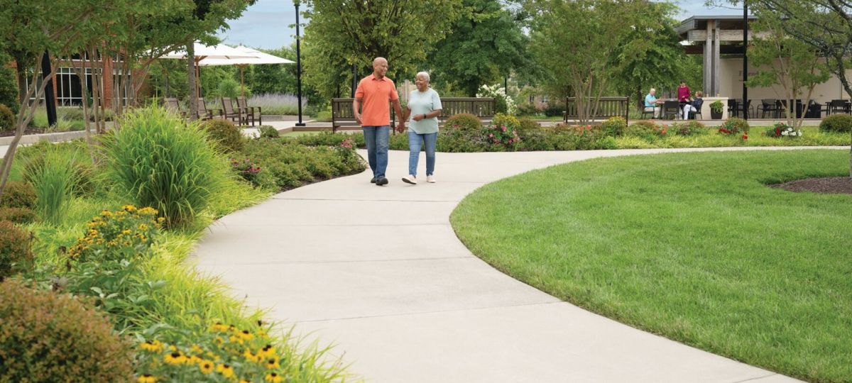 Older couple walking together along a curved path in a landscaped garden with flowers, trees, and outdoor seating areas. 