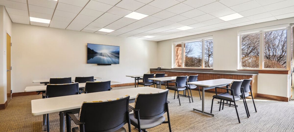 Bright meeting room with tables and chairs, large windows, and a wall-mounted TV in a modern community space.