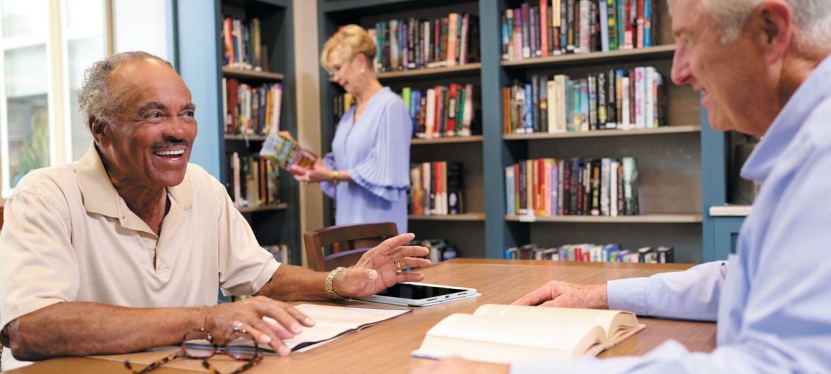 Older adults reading and conversing at a table in a cozy library with bookshelves in a senior living community.