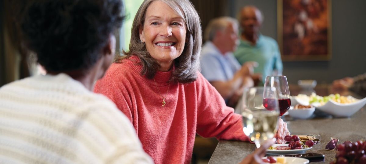 Smiling older adults enjoying drinks and conversation at a bar, with wine glasses and small plates of food in a warm, social setting.