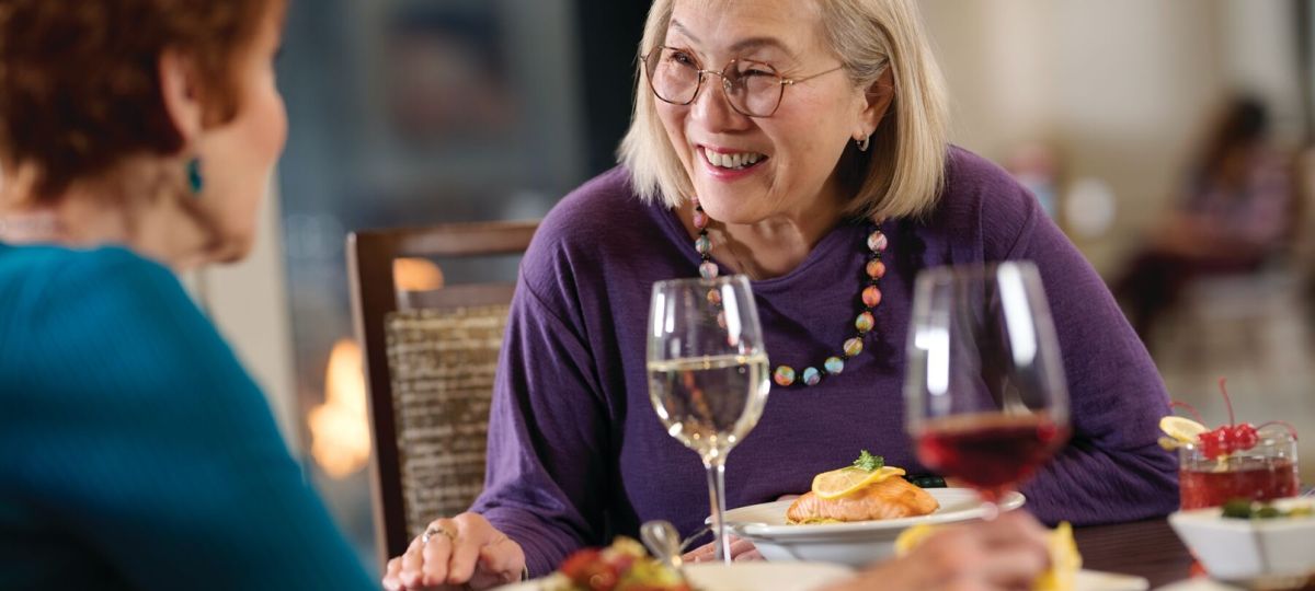 Older woman smiling and talking across a dining table with wine and plated food.