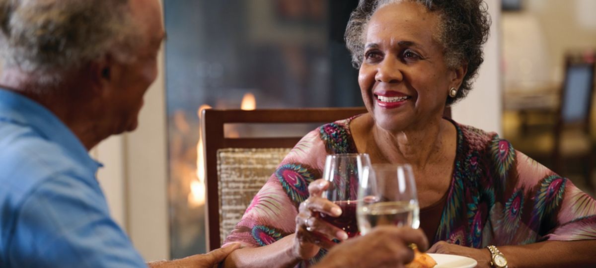 Older couple smiling and toasting with drinks at a dining table in a warm, cozy setting by a fireplace.