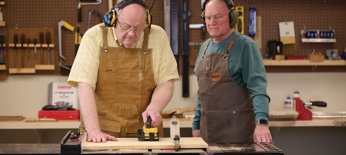 Two older men using a table saw in a workshop wearing safety gear and aprons.