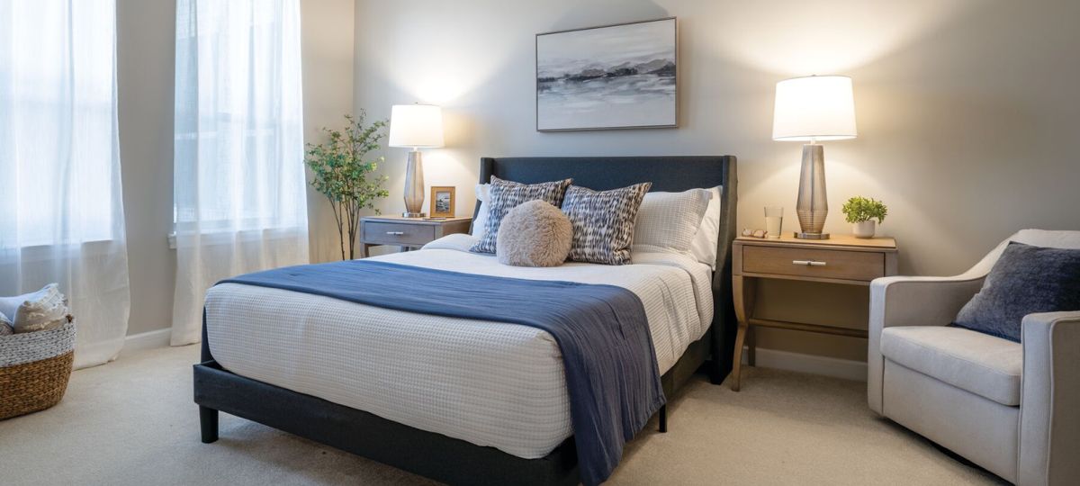 Modern staged bedroom with neutral decor, upholstered bed, bedside lamps, and soft natural light from large window.