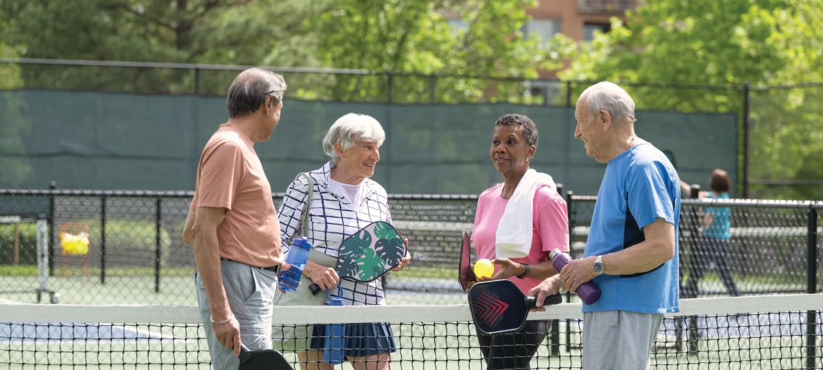 four seniors stand together talking. They are outside on a pickleball court, and are divided by the net. They are all holding pickleball rackets.