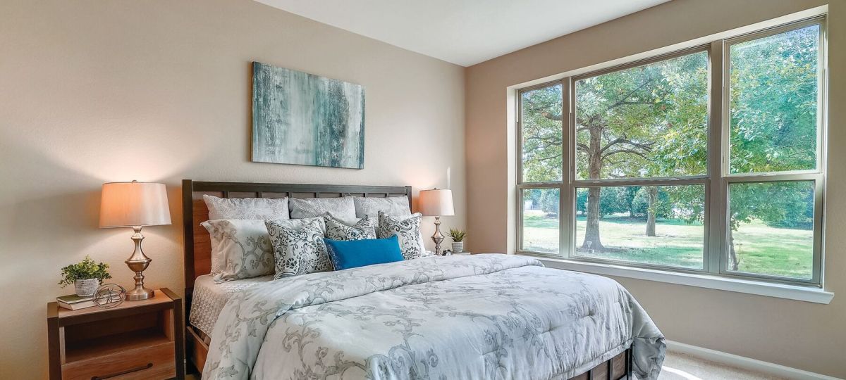 Bright bedroom with large windows, plush bedding, wood furniture, and peaceful outdoor views.