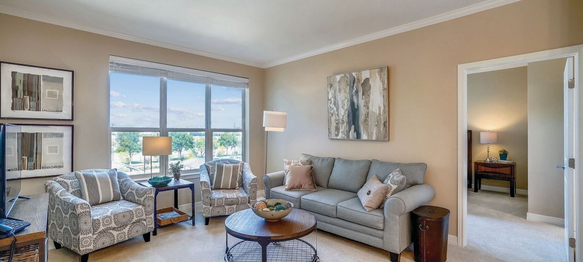 Cozy apartment living room with large window, gray sofa, patterned accent chairs, and natural light.