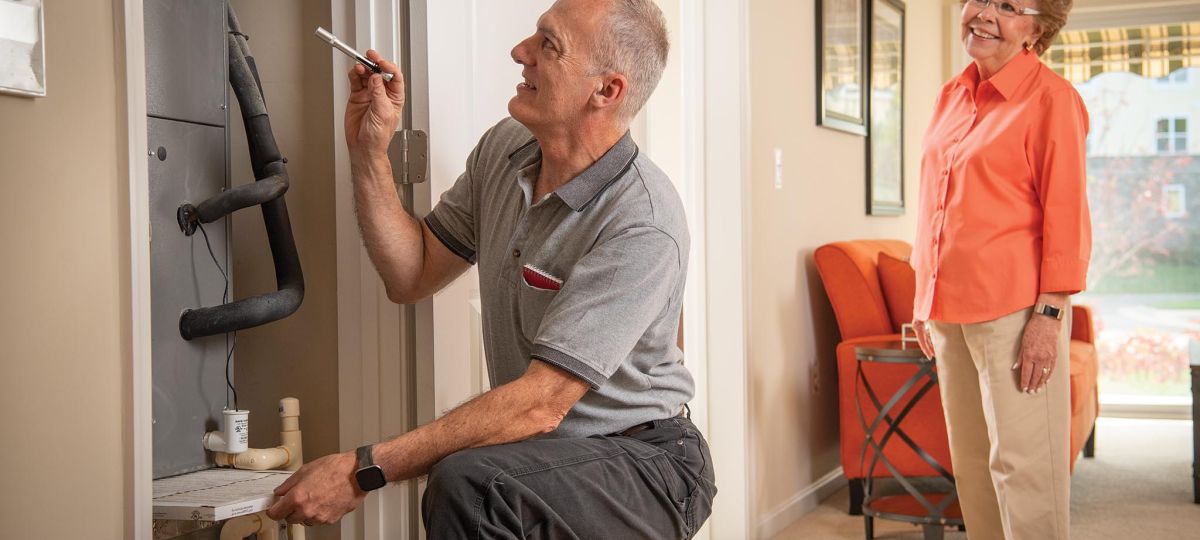 a maintenance worker observes a furnace in an apartment while a woman watches behind him