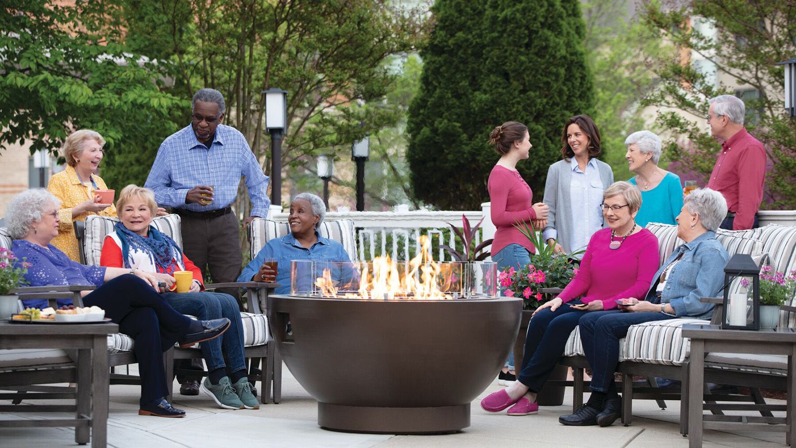 A diverse group of Riderwood residents gather around a fire pit on a community patio. 