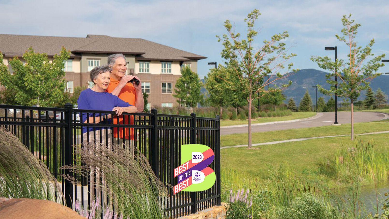  An older man and woman stand together in a gazebo overlooking a garden. The man has binoculars. Both are smiling and appear happy in each other's company. Mountains are in the background.