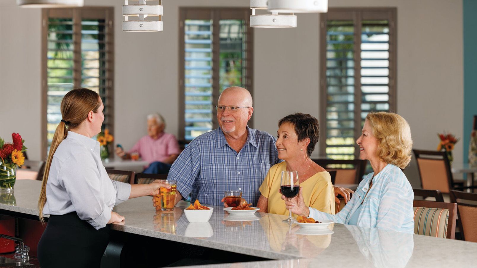 Server hands drinks to three smiling older adults sitting at a bar with snacks in a bright, modern dining area.