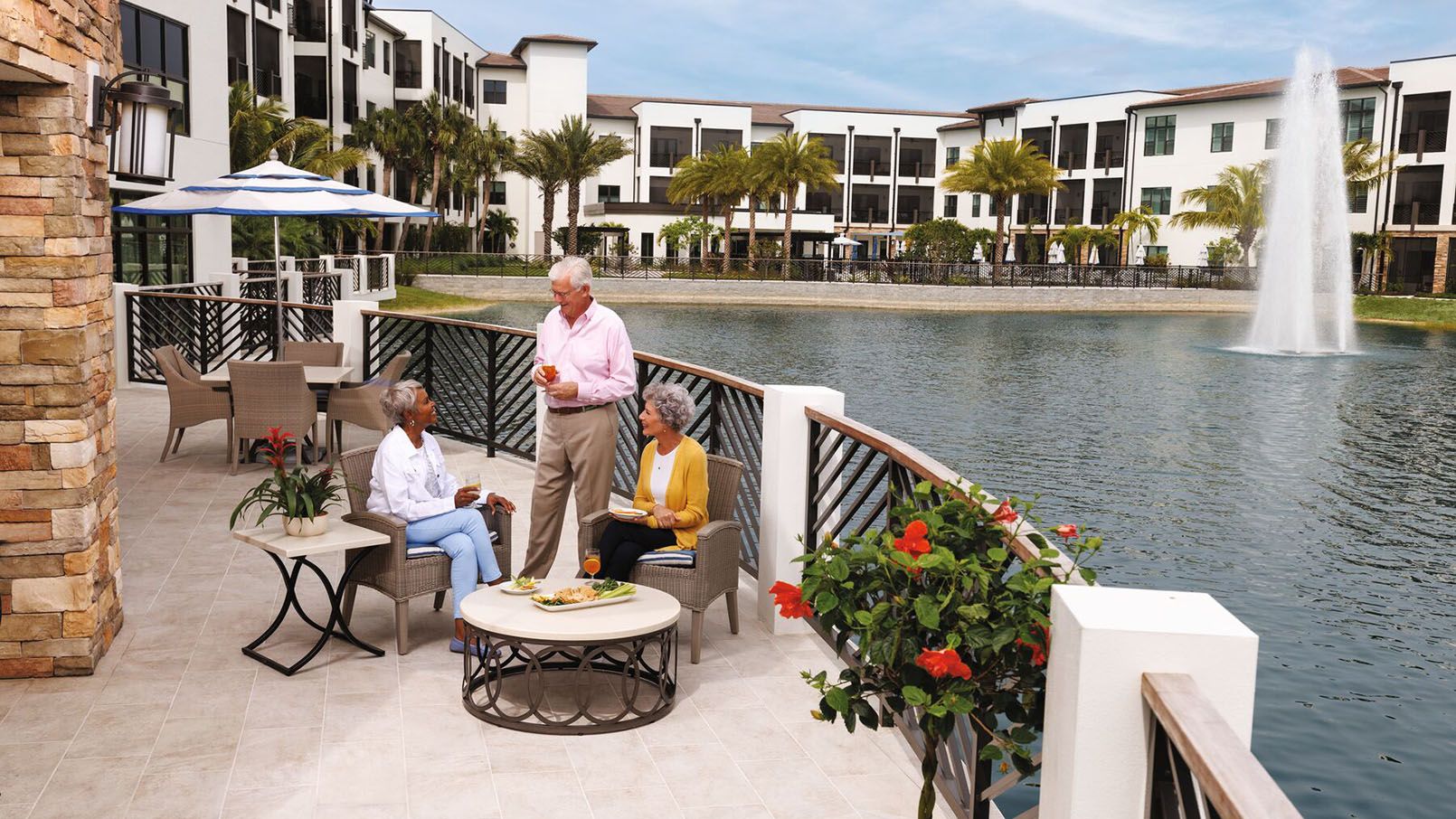 Three seniors enjoying drinks and food on a lakeside patio with a fountain and modern apartment buildings in the background.