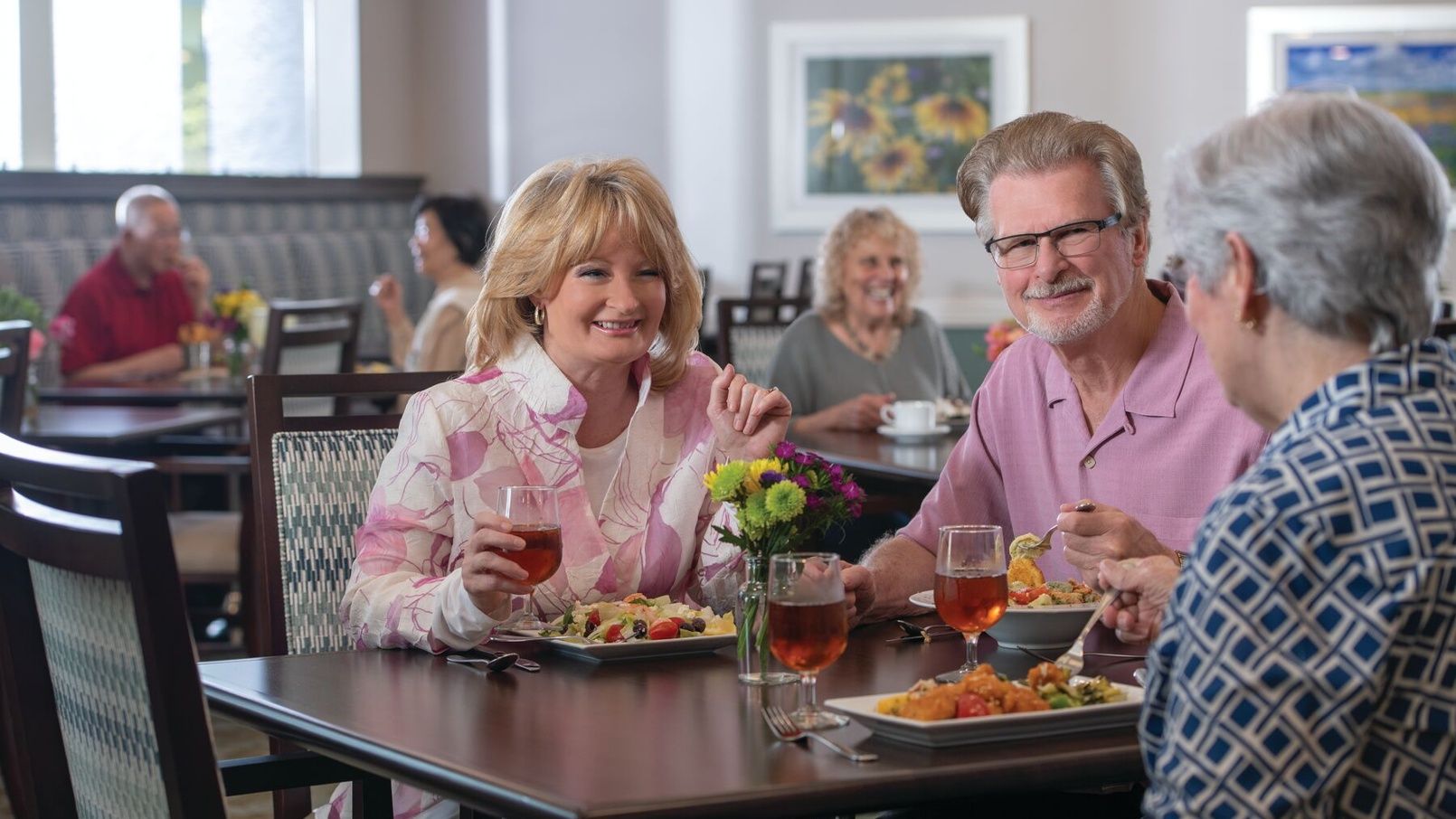 Older woman smiling and talking across a dining table with wine and plated food.