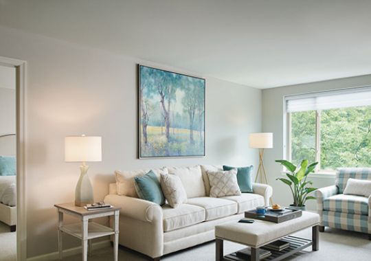 Bright and modern living room with neutral tones, featuring a beige sofa, wooden coffee table, accent chairs, and a large window with sheer curtains.
