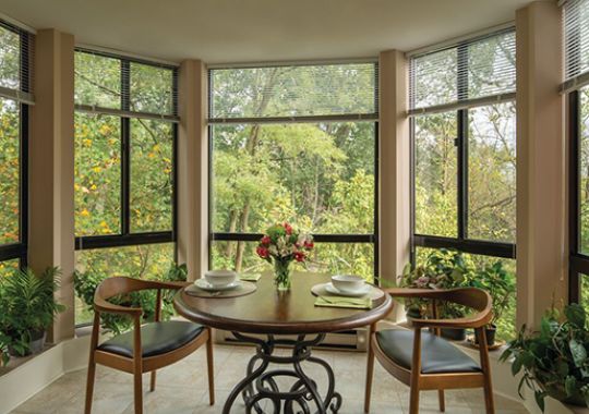 Cozy dining nook with a round wooden table, two mid-century modern chairs, and large windows overlooking lush greenery.