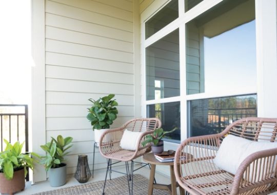 Cozy outdoor balcony with two wicker chairs, a small table, and potted plants, creating a relaxing seating area with natural light.