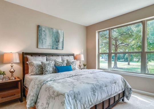 Bright bedroom with large windows, plush bedding, wood furniture, and peaceful outdoor views.