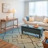 A bright and airy senior apartment living room with a large window, comfortable seating arranged for conversation, and tasteful decor creating a warm, inviting space.