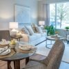 A stylish, living room with modern vinyl flooring and featuring a couch with decorative pillows and a small circular breakfast table with four cushy chairs. A cozy armchair is positioned near a large window with a scenic view of greenery.