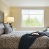 A stylish bedroom featuring a large bed with an intricate patterned headboard, matching bedding, and decorative pillows. Two elegant table lamps sit on wooden nightstands, while a cozy armchair is positioned near a window with a scenic view of greenery.