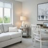A cozy home office and sitting area with a light-colored loveseat adorned with soft pillows, positioned by a large window overlooking lush greenery. A rustic wooden desk with a distressed finish holds a laptop, books, and a potted plant, accompanied by a matching chair with a cushioned seat.