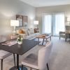Modern living and dining area with neutral tones, featuring a square dining table with plush chairs, a cozy beige sofa, contemporary wall art, and a TV mounted on the wall.