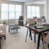 Elegant open-concept living and dining room with large windows, beige carpet, a light gray sectional sofa, a dark wood dining table with cushioned chairs, and modern decor elements.