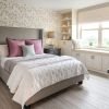 Elegant bedroom with a gray upholstered bed, white bedding, mauve and sage pillows, floral wallpaper, and dark wood nightstands. A built-in window seat with storage and a sleek desk add charm and functionality.