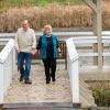 Senior couple holding hands and walking together on a wooden dock by a pond, surrounded by autumn foliage and reeds, with a fountain in the background.