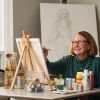 Older woman painting on a canvas at an easel, surrounded by art supplies in a bright, creative studio space. 