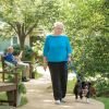 Older woman walking a dog along a garden path in an Erickson Senior Living community courtyard, with another resident seated on a bench reading nearby.
