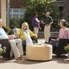 Group of older adults relaxing on an outdoor patio, sitting and standing together while enjoying drinks and conversation in a landscaped courtyard setting at an Erickson Senior Living community. 