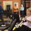 Older adults exercising in a senior living fitness center, including a woman using a rowing machine and a man walking on a treadmill, with staff assisting in the background.