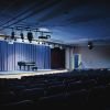 Empty auditorium with rows of seats facing a softly lit stage featuring a grand piano and closed curtains.
