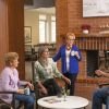Four older adults sitting and chatting comfortably around a table by a fireplace in a cozy community lounge with bookshelves and warm decor.