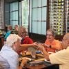 Group of older adults dining together at a restaurant table, smiling and toasting with drinks beside a wall of wine bottles.
