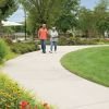 Older couple walking together along a curved path in a landscaped garden with flowers, trees, and outdoor seating areas. 