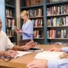 Older adults reading and conversing at a table in a cozy library with bookshelves in a senior living community.