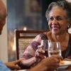 Older couple smiling and toasting with drinks at a dining table in a warm, cozy setting by a fireplace.
